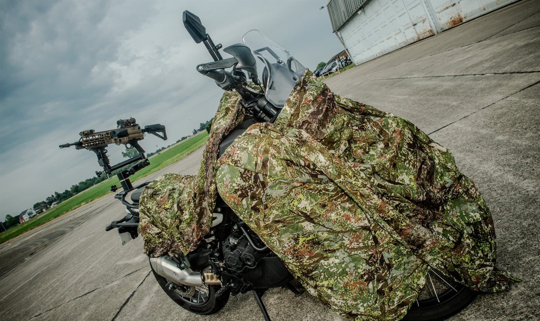 Motorcycle under a camouflage net, with an FN Evolys light machine gun on its mount standing behind it on the apron